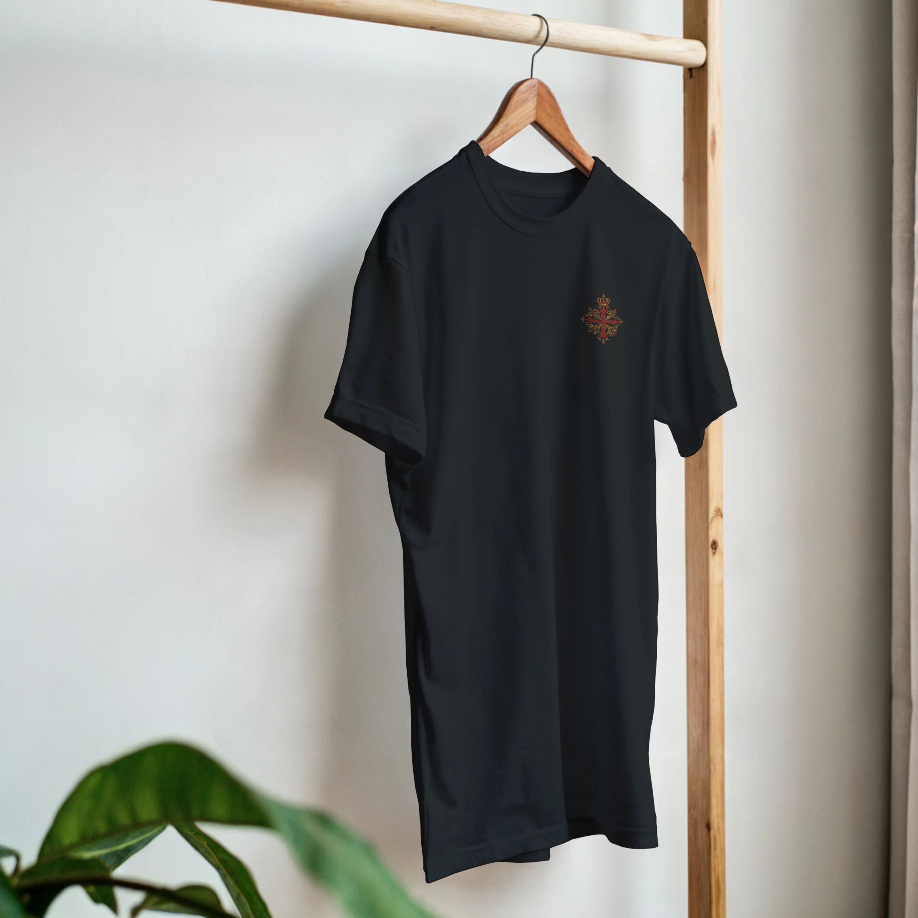Camiseta negra con cruz coronada, diseño inspirado en el legado español, colgada en perchero de madera.